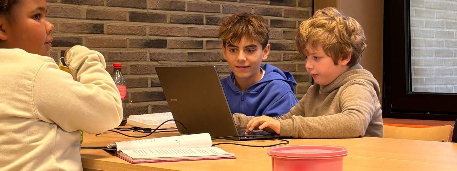 Drie kinderen van de plusklas aan een tafel met een laptop en een open boek; twee jongens werken samen op de laptop.