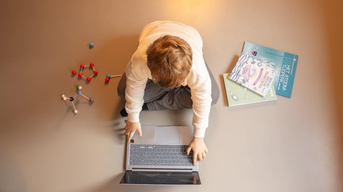 Van bovenaf gezien zit een jongen op de grond te typen op een laptop, met molecuulmodellen en boeken over natuurkunde rondom zich.