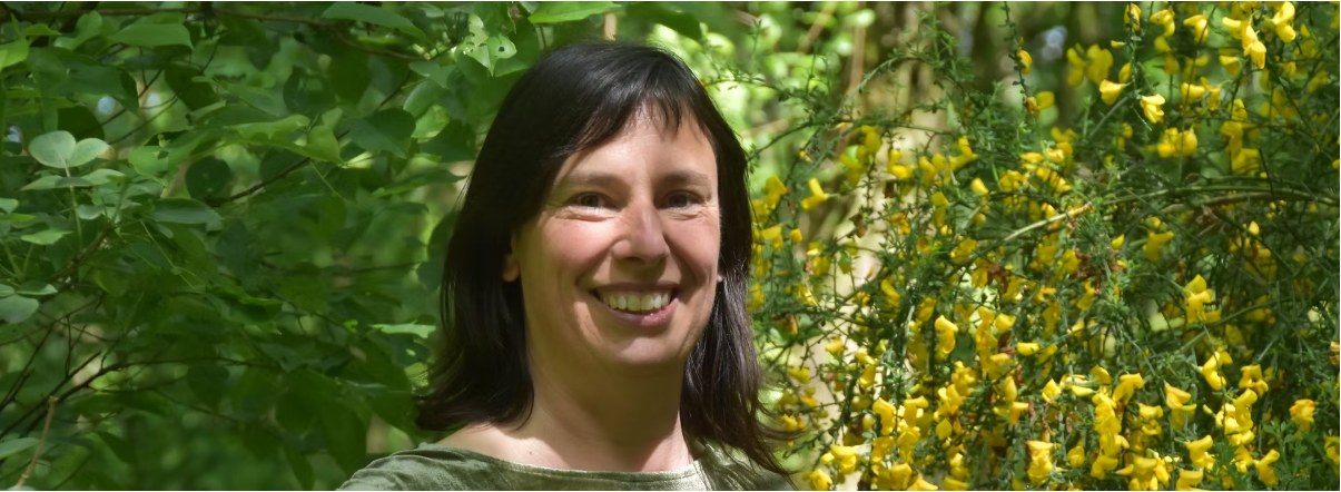 Katrien Volckaert, lachend, voor groen struikgewas met gele bloemen.