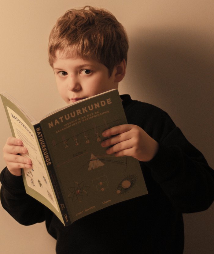 Een jongen houdt een visuele gids over natuurkunde open en kijkt over het boek heen.