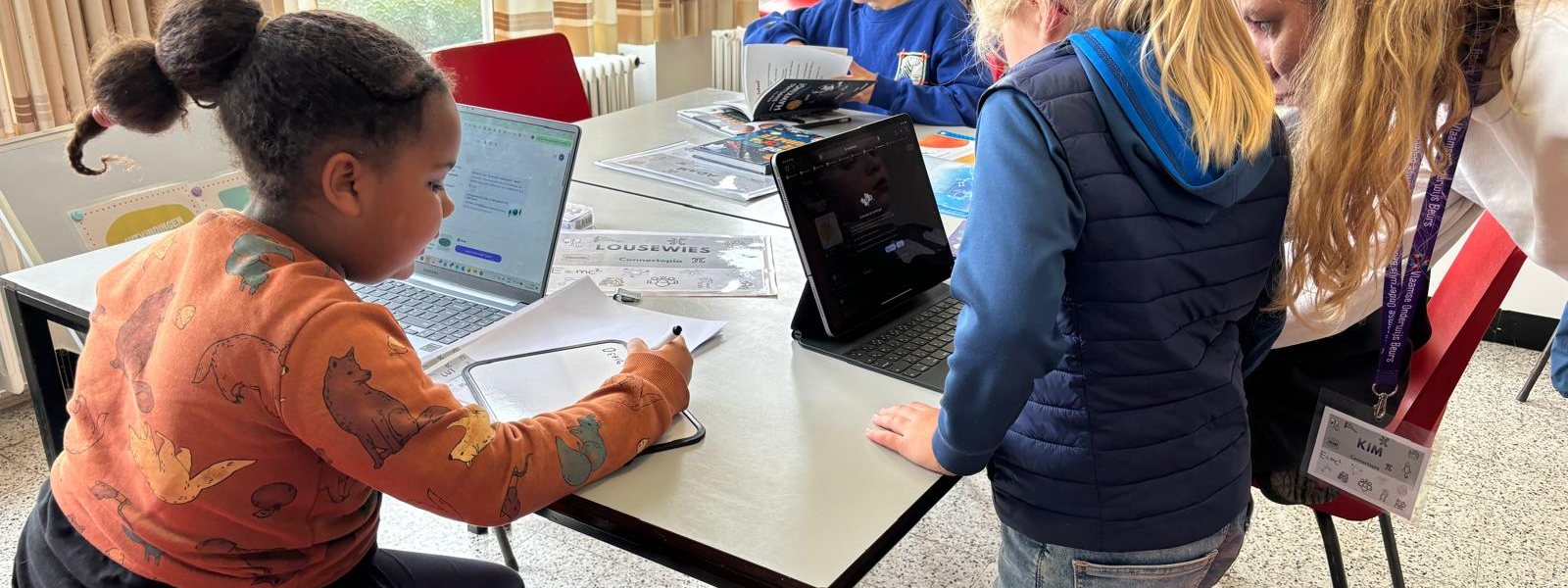 Kinderen werken aan een lange tafel met laptops en boeken, een begeleidster kijkt mee.
