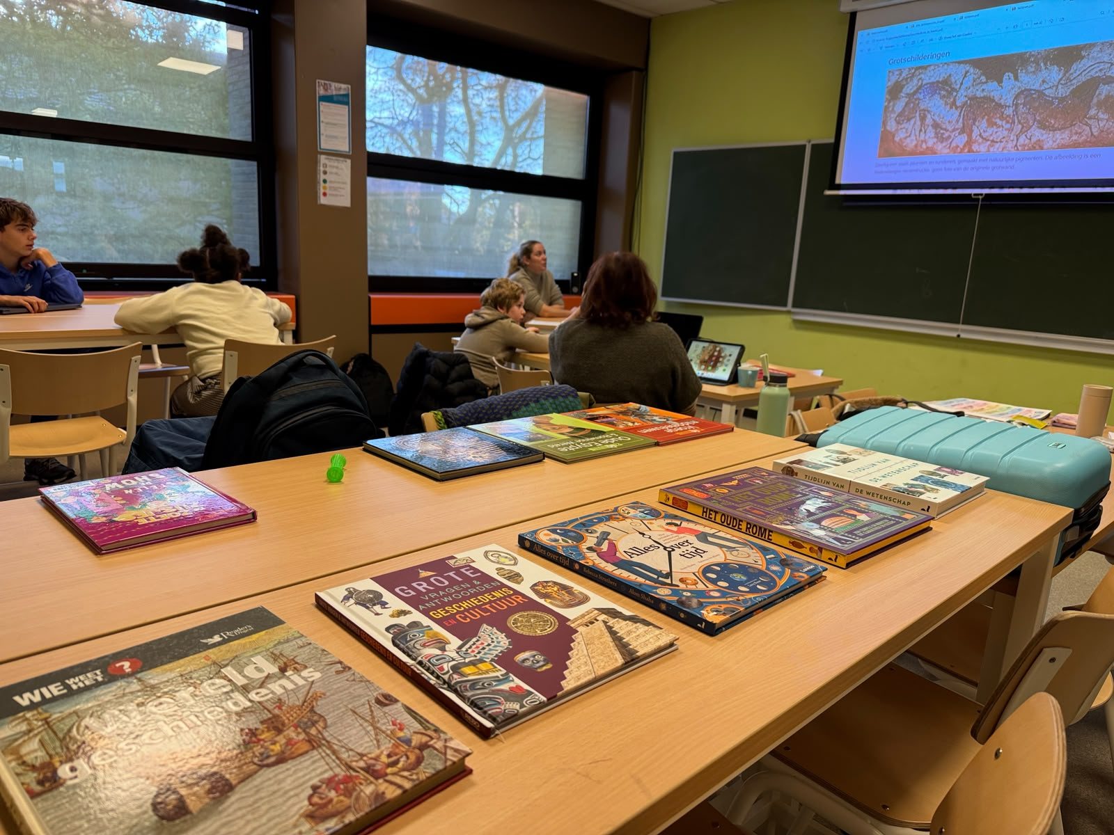 Klaslokaal met geschiedenisboeken op de tafels vooraan en grotschilderingen op het grote scherm