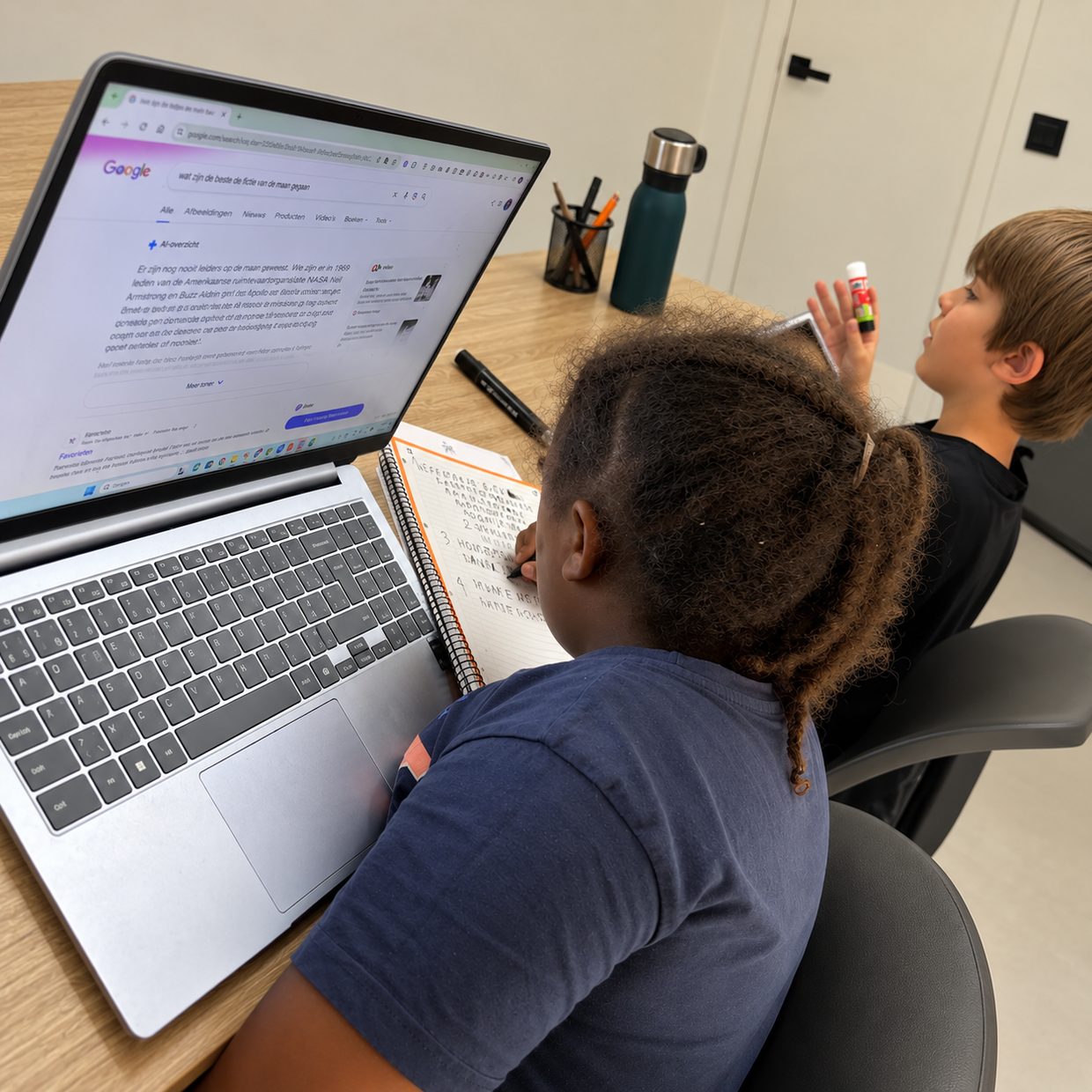 Twee kinderen zoeken iets op een laptop op en schrijven mee in een schrift